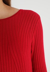 Primo piano di una persona che indossa un maglione rosso a coste con motivi diagonali e verticali su uno sfondo neutro.