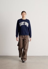 Tengerészkék pulóver "GANT 1949" felirattal, laza barna nadrággal párosítva. Simább anyag és egyszerű dizájn. Egyszínű háttér.