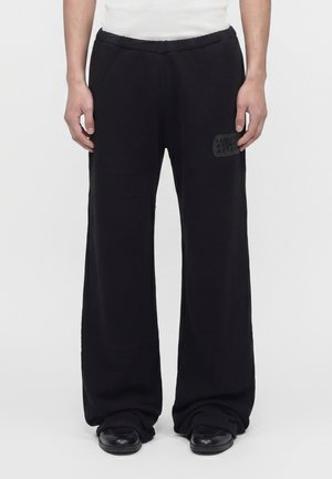 Vue de face d'une personne portant un pantalon noir à jambes larges avec un patch sur la cuisse gauche, des chaussures noires et une chemise blanche, debout sur un sol blanc.