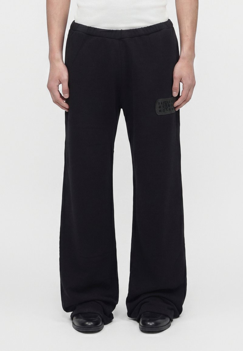 Vue de face d'une personne portant un pantalon noir à jambes larges avec un patch sur la cuisse gauche, des chaussures noires et une chemise blanche, debout sur un sol blanc.