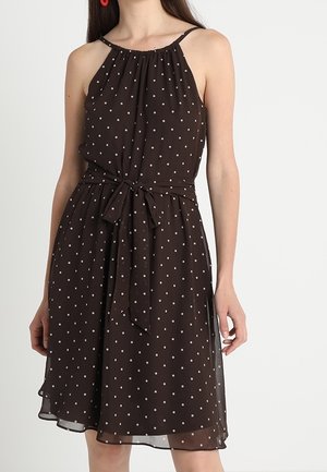 Czarna sukienka bez rękawów w białe kropki, z marszczonym dekoltem i wiązaniem w talii, noszona przez kobietę z długimi brązowymi włosami.