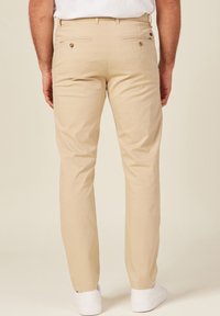 Pantalones de algodón beige con corte recto, que cuentan con dos bolsillos traseros con cierres de botón y una textura sutil, combinados con zapatos blancos.
