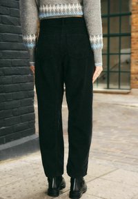 Pantaloni a vita alta in corduroy neri con design a gamba dritta e tasche posteriori. Indossati con un maglione grigio a fantasia e stivaletti neri.