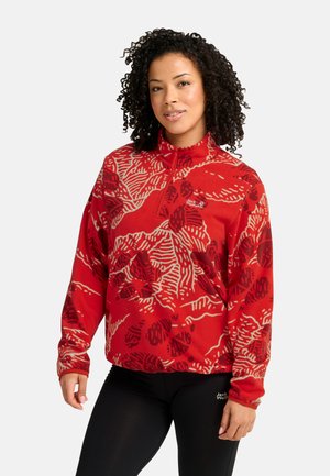 Donna con capelli ricci che indossa un maglione in pile rosso a motivo e leggings neri, in piedi contro uno sfondo semplice.