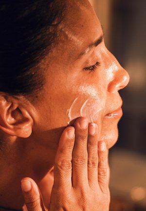 Nahaufnahme einer Person, die einen cremigen, weißen Gesichtsreiniger auf ihre Wange aufträgt; die Haut wirkt glatt und leicht nass mit einem natürlichen Glanz.