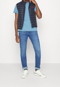 Marineblaues gepolstertes Weste über einem hellblauen T-Shirt, kombiniert mit blauen Jeans und weißen Sneakern. Die Weste hat ein Logo und hochgekrempelte Ärmel.