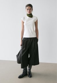 Femme portant un haut blanc à manches courtes, un pantalon large noir raccourci, des bottes noires, un foulard vert autour du cou et tenant un sac à main noir.