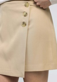 Jupe A-line beige avec une fermeture sur le côté, composée de trois boutons transparents et d'un tissu lisse et texturé pour un look soigné.