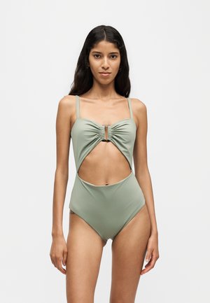 Femme aux longs cheveux foncés portant un maillot de bain une pièce vert clair avec découpes, debout devant un fond blanc uni.