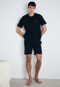 Svart bomull t-skjorte med lite logo og matchende shorts. Enkel, uformell design med en avslappet passform. Barfot på et lysfarget teppe.