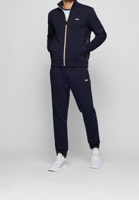 Marinträningsdräkt med zip-jacka och byxor med elastiskt midjeband. Rosa dragkedja som accent. Modellen bär vita sneakers för en sportig look.