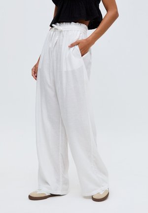 Frau trägt lockere weiße Hosen mit Kordelzug und seitlichen Taschen, kombiniert mit beigen und weißen Sneakern und einem schwarzen bauchfreien Oberteil.