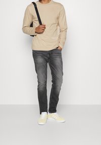 Mężczyzna w beżowej koszuli z długim rękawem, szarych dżinsach, bladożółtych trampkach, niosący czarną torbę na ramię, z ręką w kieszeni na jednolitym tle.