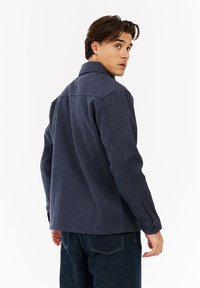 Jonge man die over zijn schouder kijkt, gekleed in een donkerblauwe jas en een donkere spijkerbroek, tegen een effen witte achtergrond.