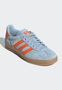 Light blue suede sneakers with orange stripes, rubber sole, and textured accents on the tongue. Features "Gazelle" logo in yellow.