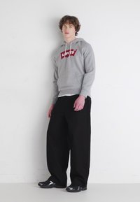 Grijze hoodie met een rood Levi's logo, gecombineerd met losse zwarte broek. Model staat tegen een witte muur, draagt zwarte sportschoenen.