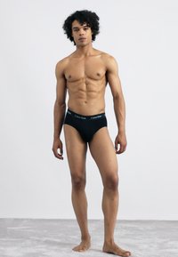 Čierne bavlnené slipy s elastickým pásom so značkou "Calvin Klein". Model stojí vzpriamene a predvádza priliehavý dizajn a hladkú textúru.