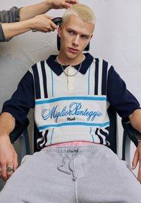 Polohemd in Creme, Marineblau und Blau mit gestreiftem Muster und gesticktem Text. Graue Jogginghose, silberne Kettenhalskette und ein Ring.