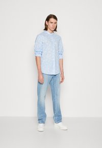 Camicia azzurro chiaro con motivo a foglie bianche, maniche arrotolate, abbinata a jeans di denim chiaro e sneakers bianche, in piedi contro uno sfondo neutro.