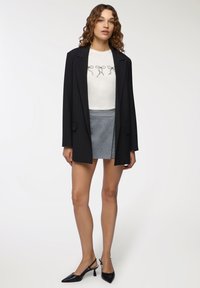 Blazer lungo nero, camicia bianca con grafica a fiocco nero, mini gonna grigia, décolleté neri con slingback. Tessuto morbido, vestibilità su misura, design minimalista.