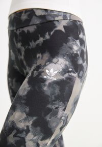 Čierne a sivé legíny s batikovaným vzorom. Vyrobené z elastického materiálu s bielym logom Adidas blízko bokov.