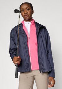 Granatowa wodoodporna kurtka z ściągaczem u dołu, noszona na różowym topie na suwak. Modelka trzyma kij do golfa, stoi na jasnym tle.