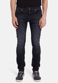 Jeans in denim blu scuro con una vestibilità slim, chiusura a bottoni, cinque tasche e un sottile dettaglio di whiskering. Abbinati a sneakers nere, che mostrano una texture stratificata.
