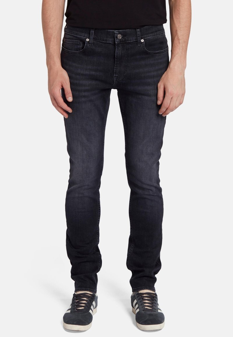 Jeans in denim blu scuro con una vestibilità slim, chiusura a bottoni, cinque tasche e un sottile dettaglio di whiskering. Abbinati a sneakers nere, che mostrano una texture stratificata.
