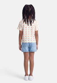 Παιδί με μακριά πλεξούδα, φοράει κοντομάνικο πουκάμισο με σχέδιο κεράσια, τζιν σορτς και λευκά αθλητικά, στέκεται με την πλάτη γυρισμένη.