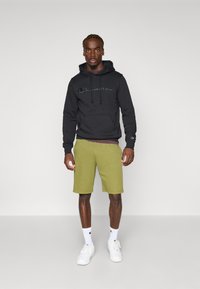 Sweat-shirt à capuche noir avec le logo "Champion", shorts verts et baskets blanches. Le modèle se tient sur un fond neutre. Les chaussettes sont blanches avec logo.