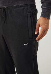 Zwarte fleece joggers met een elastische tailleband met een trekkoord, zijzakken en een wit Nike swoosh-logo op de linker dij.