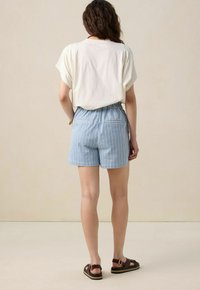 Haut blanc à manches courtes, shorts rayés bleu clair et blanc avec une taille élastique, et sandales brun foncé avec des boucles.