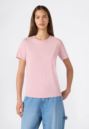 Mujer que lleva una camiseta lisa de manga corta color rosa claro y vaqueros azul claro, de pie frente a un fondo blanco.