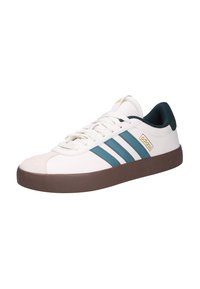 Zapatillas blancas con tres franjas en teal, una puntera de ante beige y una suela de goma marrón. Presentan un cuello acolchado en verde oscuro.