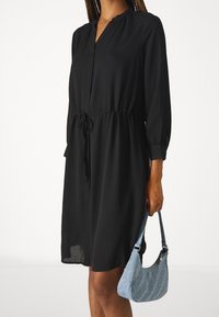 Svart långärmad klänning med rund halsringning, åtsittande midja och ett lätt tyg; matchad med en blå handväska med dekorativa detaljer.