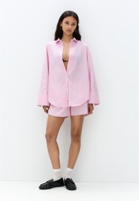 Camicia a maniche lunghe rosa e bianca a righe, vestibilità morbida, con orlo curvo, abbinata a pantaloni corti coordinati e mocassini neri con calze bianche.