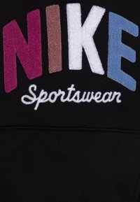 NIKE Sportswear-logotyp med stora texturerade bokstäver i rosa, brun, vit och blå på svart tygbakgrund.