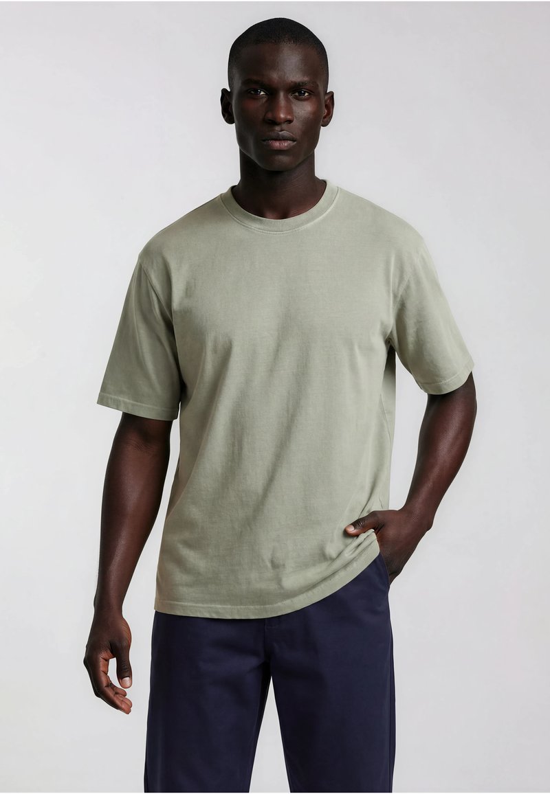 Homme portant un t-shirt vert clair uni à manches courtes et un pantalon bleu foncé, debout avec une main dans la poche devant un fond uni.
