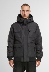 Parka călduroasă și izolată, de culoare neagră, cu glugă, mai multe buzunare frontale, manșete cu nervuri și închidere cu fermoar. Textură de țesătură netedă și mată.