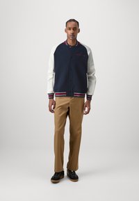 Chaqueta varsity azul marino y blanca con acentos rojos, cierre de botones, cuello y puños acanalados, combinada con pantalones beige y zapatillas negras.