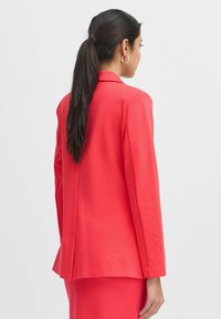 Rode op maat gemaakte blazer met een gladde textuur, notch revers, lange mouwen en een achteropening, gedragen over een bijpassende rok.