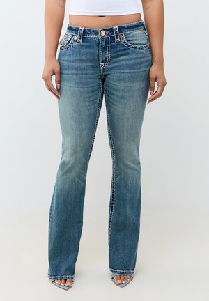 Vue de face d'une personne portant un jean bootcut bleu délavé et des talons ouverts bleu clair, un top blanc court, debout devant un fond uni.