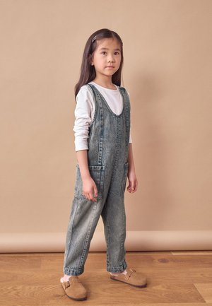 Denim-Latzhose mit zwei Vorderaschen, hellblauer Farbe, entspannter Schnitt, getragen über einem weißen Langarmhemd, kombiniert mit braunen Holzclogs.