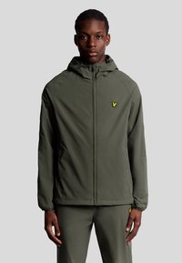 Giacca leggera verde oliva con zip frontale, cappuccio regolabile e un piccolo logo giallo sul lato sinistro del petto. Tessuto liscio, design minimale.