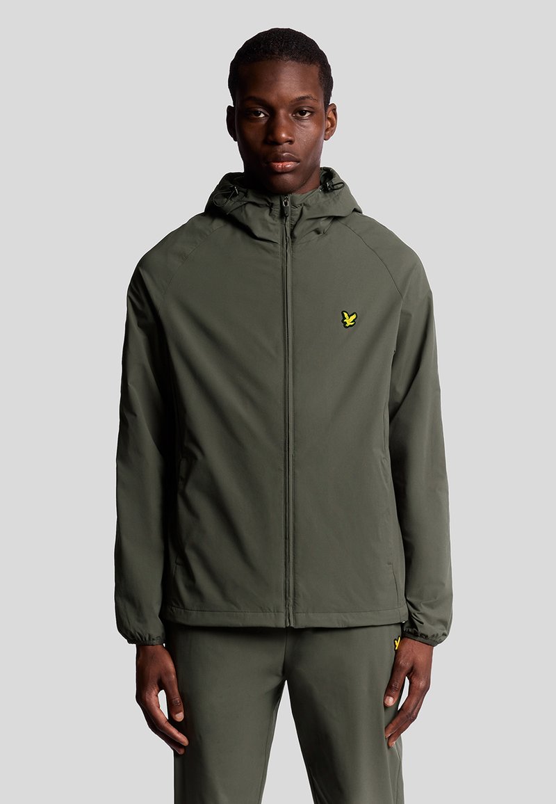 Giacca leggera verde oliva con zip frontale, cappuccio regolabile e un piccolo logo giallo sul lato sinistro del petto. Tessuto liscio, design minimale.