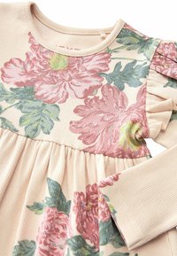 Robe bébé à manches longues avec imprimé floral rose, détail volant sur les épaules et tissu texturé côtelé sur fond beige clair.