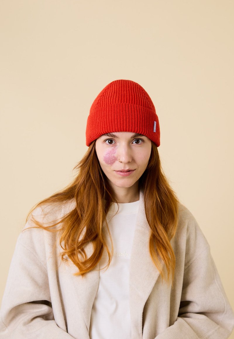 Red knitted beanie with ribbed texture, featuring a small blue tag. Complemented by a beige coat and light sweater underneath.