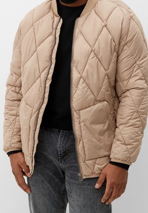 Homme portant une veste matelassée beige sur une chemise noire et un jean gris foncé, avec la veste partiellement ouverte et les mains détendues le long du corps.