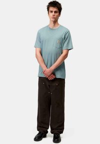 Camiseta de algodón azul claro con un bolsillo en el pecho, combinada con pantalones holgados de tono marrón oscuro que presentan costuras visibles y detalles metálicos.