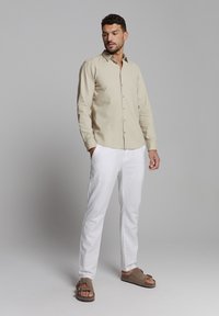 Beige overhemd met knopen, witte broek, bruine sandalen. Het overhemd heeft een zachte textuur en een kraag, terwijl de broek op maat is gemaakt met een rechte snit.